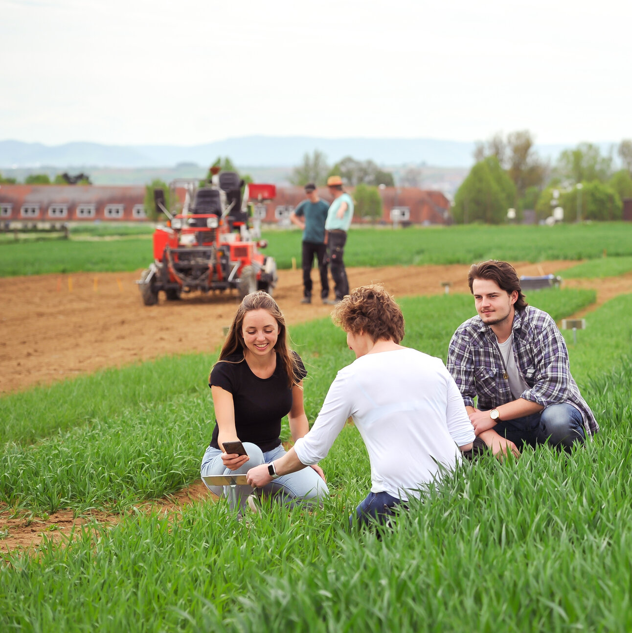 Foto: Eine Gruppe von Personen sitzt auf einem grünen Feld. Im Hintergrund sind landwirtschaftliche Maschinen und weitere Personen zu sehen. Die Umgebung wirkt ländlich und weitläufig.