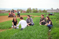 Foto: Eine Gruppe von Personen sitzt auf einem grünen Feld. Im Hintergrund sind landwirtschaftliche Maschinen und weitere Personen zu sehen. Die Umgebung wirkt ländlich und weitläufig.