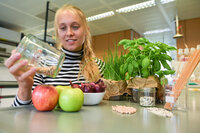 Foto: Eine Person in einem gestreiften Oberteil hält ein Glas mit Samen. Auf dem Tisch liegen Äpfel, Zwiebeln, Kräuterpflanzen und weitere Samen. Im Hintergrund ist ein Labor zu sehen.