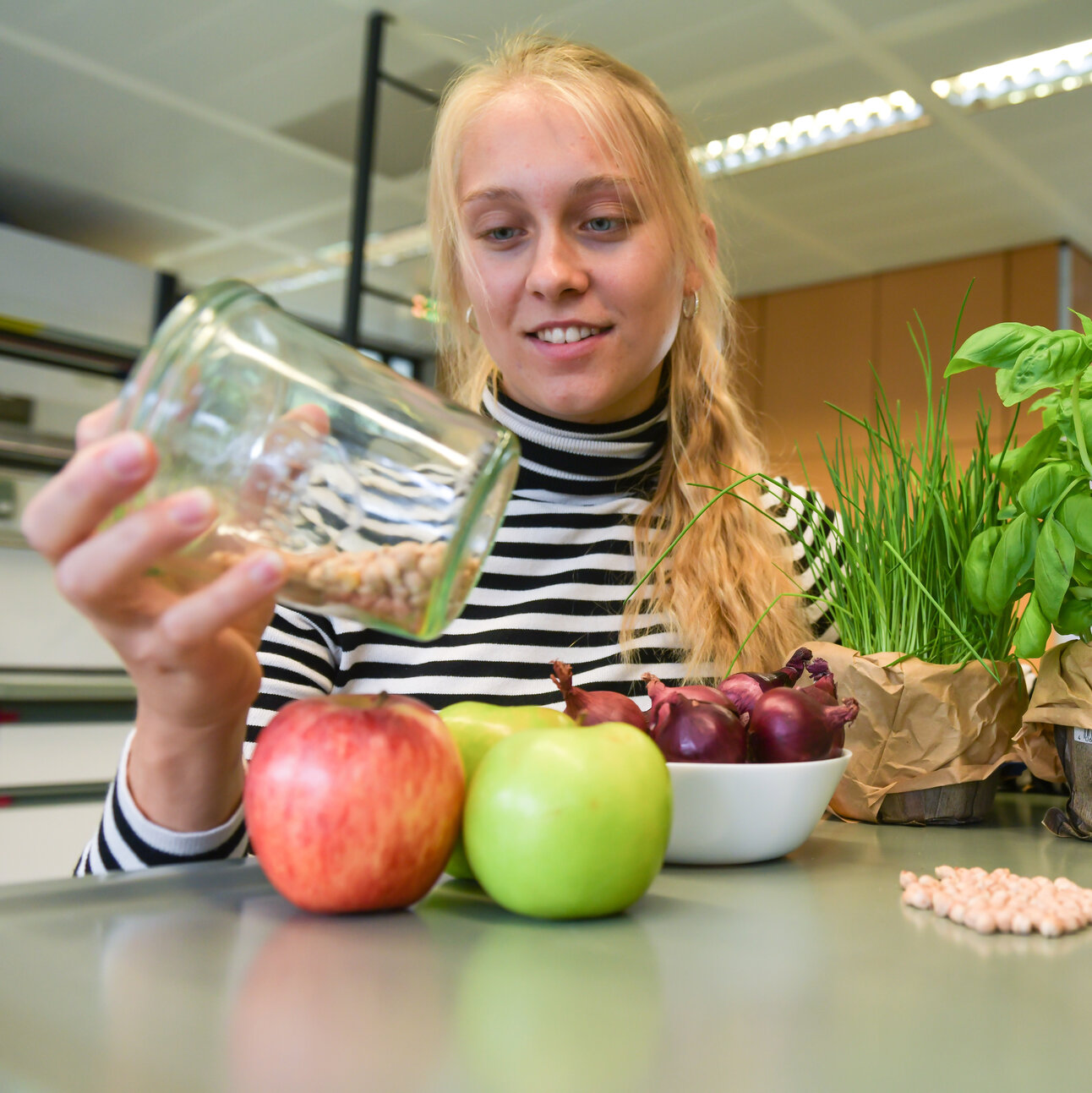 Foto: Eine Person in einem gestreiften Oberteil hält ein Glas mit Samen. Auf dem Tisch liegen Äpfel, Zwiebeln, Kräuterpflanzen und weitere Samen. Im Hintergrund ist ein Labor zu sehen.