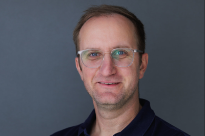 Das Porträt zeigt Heiko Breitenbach lächelnd mit verschränkten Armen vor grauem Hintergrund.