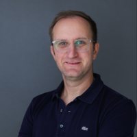 Das Porträt zeigt Heiko Breitenbach lächelnd mit verschränkten Armen vor grauem Hintergrund.