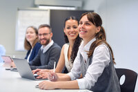 Foto: Mehrere Personen sitzen an einem Tisch in einem Büro und verwenden Laptops. Eine Person hält einen Stift. Im Hintergrund ist ein Flipchart sichtbar.