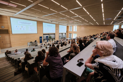 Foto: Ein großer Hörsaal mit vielen Personen, die einer Präsentation folgen. Auf der Leinwand ist der Text 'LOGIKEN NEUER' zu sehen. Der Raum ist modern gestaltet mit Holzverkleidung und großen Fenstern.