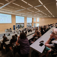 Foto: Ein großer Hörsaal mit vielen Personen, die einer Präsentation folgen. Auf der Leinwand ist der Text 'LOGIKEN NEUER' zu sehen. Der Raum ist modern gestaltet mit Holzverkleidung und großen Fenstern.