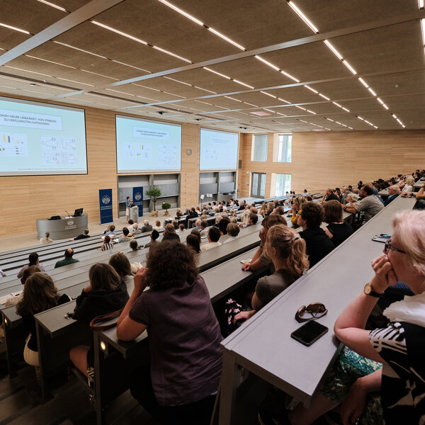 Foto: Ein großer Hörsaal mit vielen Personen, die einer Präsentation folgen. Auf der Leinwand ist der Text 'LOGIKEN NEUER' zu sehen. Der Raum ist modern gestaltet mit Holzverkleidung und großen Fenstern.