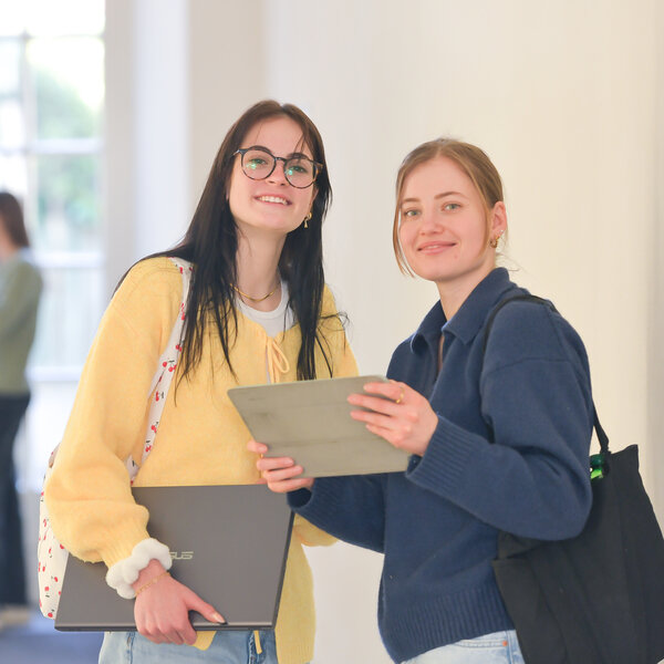 Foto: Zwei Personen stehen in einem Flur. Eine Person hält ein Tablet, die andere einen Laptop. Im Hintergrund sind zwei weitere Personen in einem Gespräch zu sehen.