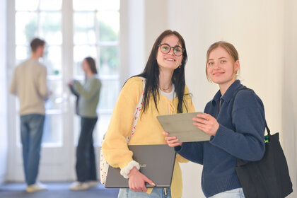 Foto: Zwei Personen stehen in einem Flur. Eine Person hält ein Tablet, die andere einen Laptop. Im Hintergrund sind zwei weitere Personen in einem Gespräch zu sehen.