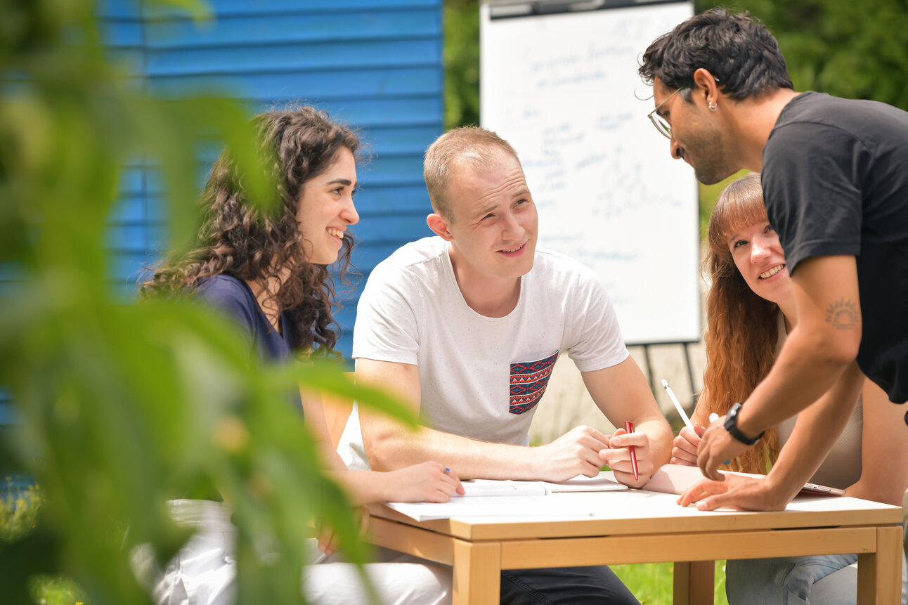 Foto: Eine Gruppe von Personen sitzt an einem Tisch im Freien und arbeitet zusammen. Im Hintergrund ist ein Flipchart mit unleserlichem Text zu sehen. Pflanzen sind im Vordergrund sichtbar.