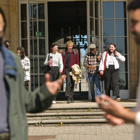 Foto: Eine Gruppe von Personen verlässt ein Gebäude durch einen Eingang mit Glastüren. Zwei Personen im Vordergrund stehen sich gegenüber und scheinen sich zu unterhalten.
