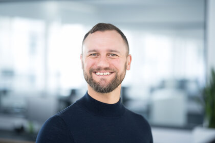 Foto: Eine Person in einem dunklen Oberteil steht in einem modernen Büro mit verschwommenem Hintergrund.