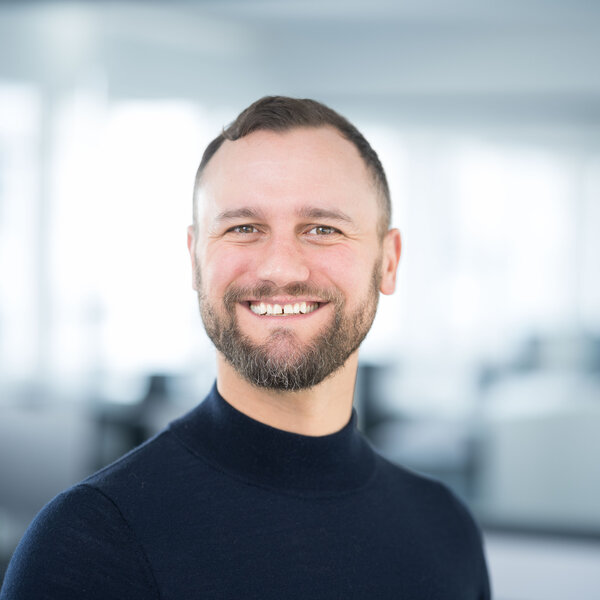 Foto: Eine Person in einem dunklen Oberteil steht in einem modernen Büro mit verschwommenem Hintergrund.