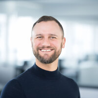 Foto: Eine Person in einem dunklen Oberteil steht in einem modernen Büro mit verschwommenem Hintergrund.