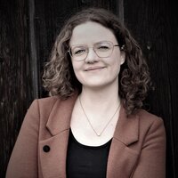Das Porträt zeigt Angela Nießer lächelnd mit verschränkten Armen vor dunklem Hintergrund.