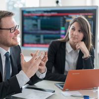 Foto: Drei Personen in Business-Kleidung sitzen an einem Tisch mit Laptops und Papieren. Im Hintergrund ist ein großer Bildschirm mit Diagrammen und Daten sichtbar.