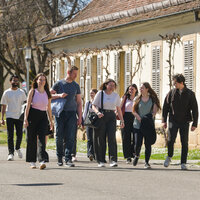 Foto: Eine Gruppe von Personen geht auf einer sonnigen Straße entlang eines Gebäudes mit Fensterläden. Im Hintergrund sind Bäume und ein Verkehrsschild mit der Aufschrift '30' zu sehen.