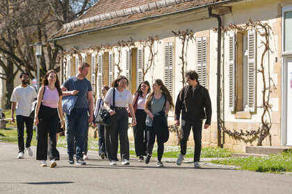 Foto: Eine Gruppe von Personen geht auf einer sonnigen Straße entlang eines Gebäudes mit Fensterläden. Im Hintergrund sind Bäume und ein Verkehrsschild mit der Aufschrift '30' zu sehen.