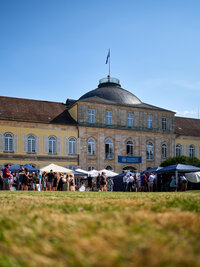 Foto: Eine Gruppe von Personen steht vor dem Schloss Hohenheim auf einem Rasen. Im Hintergrund ist das historische Gebäude mit einer Kuppel und mehreren Fenstern zu sehen. Ein Banner mit der Aufschrift 'Universität Hohenheim' hängt an der Fassade.