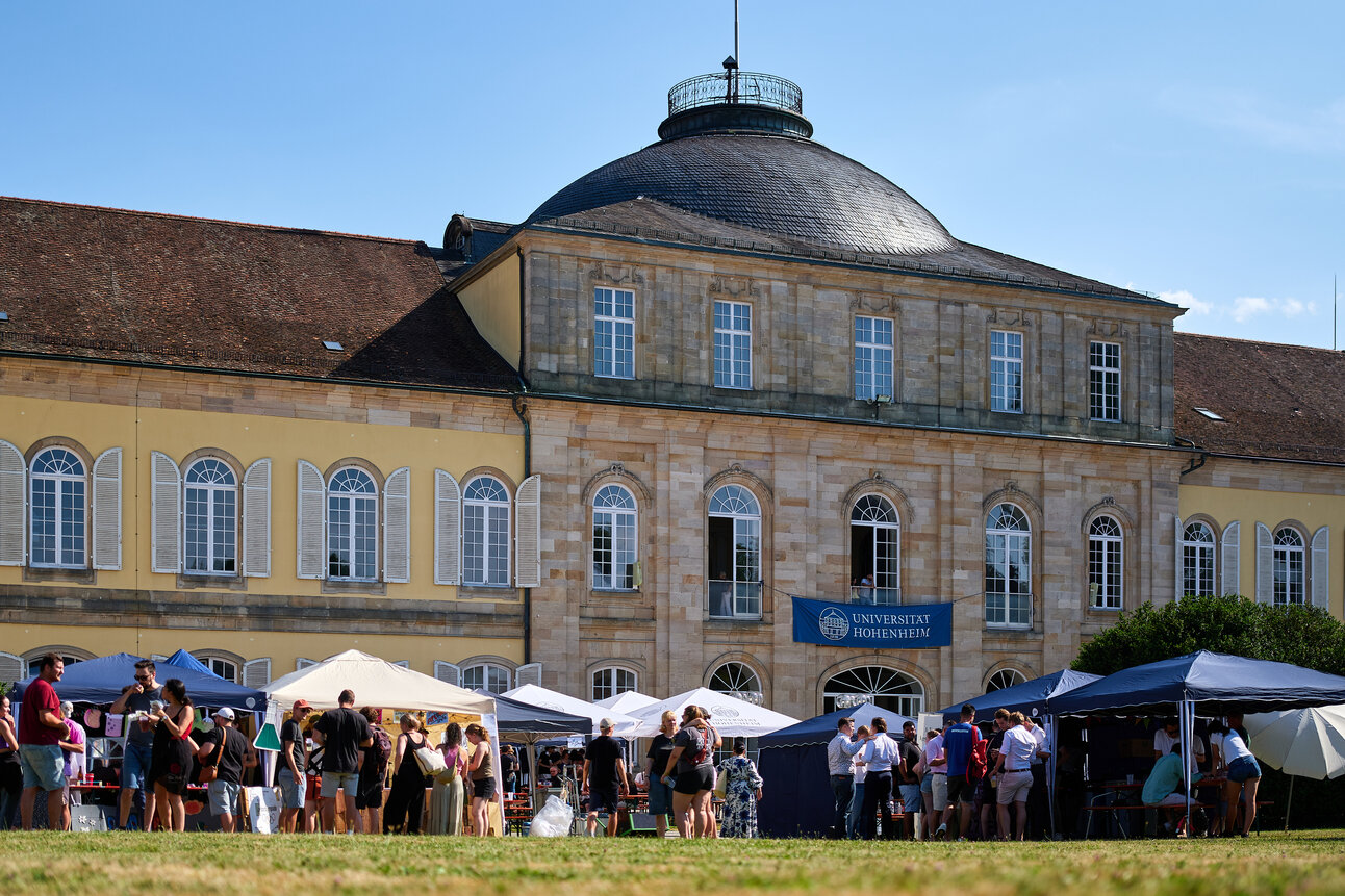 Foto: Eine Gruppe von Personen steht vor dem Schloss Hohenheim auf einem Rasen. Im Hintergrund ist das historische Gebäude mit einer Kuppel und mehreren Fenstern zu sehen. Ein Banner mit der Aufschrift 'Universität Hohenheim' hängt an der Fassade.