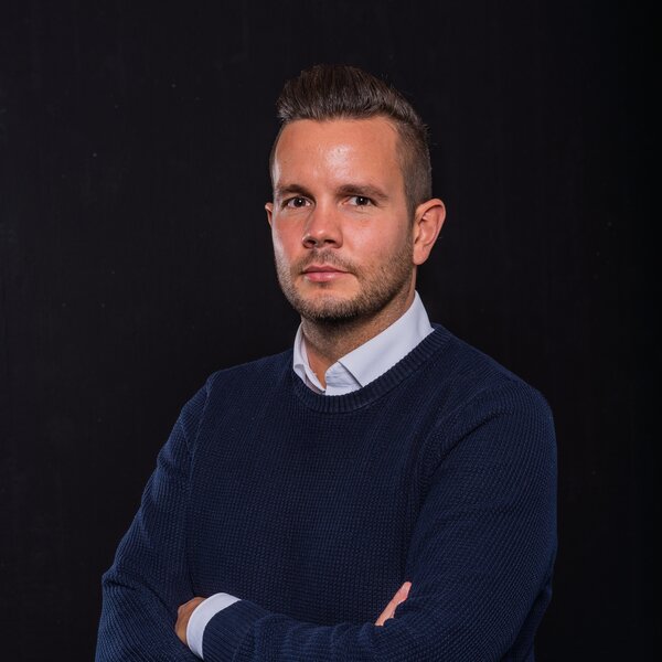 Das Porträt zeigt Daniel Schneider mit verschränkten Armen vor dunklem Hintergrund.