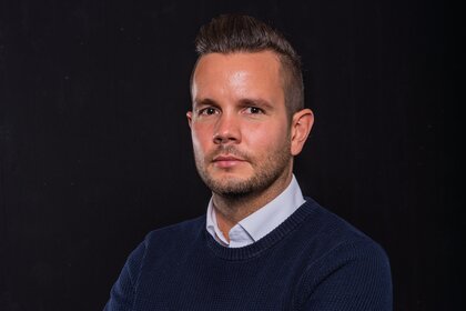 Das Porträt zeigt Daniel Schneider mit verschränkten Armen vor dunklem Hintergrund.