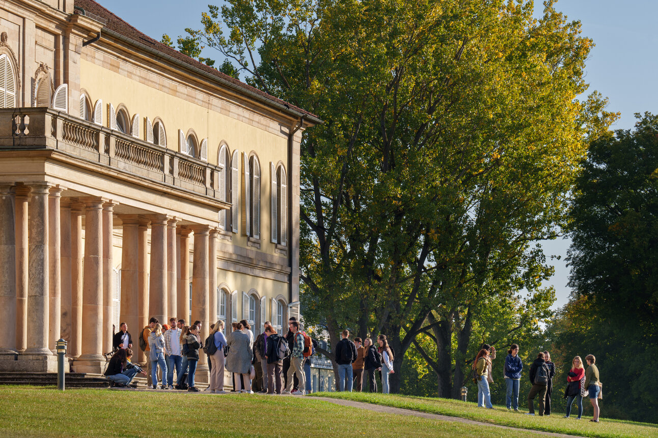 Foto: Eine Gruppe von Personen steht vor dem Schloss Hohenheim. Das Gebäude zeigt klassische Architektur mit Säulen und vielen Fenstern. Im Hintergrund sind große Bäume und ein klarer Himmel sichtbar.