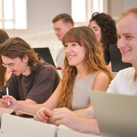 Foto: Eine Gruppe von Personen sitzt in einem Hörsaal und macht Notizen auf Laptops und mit Stiften. Die Umgebung wirkt akademisch und konzentriert.