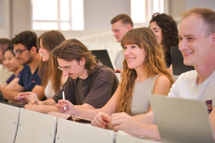 Foto: Eine Gruppe von Personen sitzt in einem Hörsaal und macht Notizen auf Laptops und mit Stiften. Die Umgebung wirkt akademisch und konzentriert.