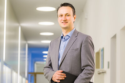 Das Porträt zeigt Prof. Dr. Ronny Baierl lächelnd mit einer Aktentasche unter dem Arm im Flur eines Gebäudes.