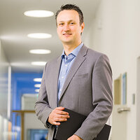 Das Porträt zeigt Prof. Dr. Ronny Baierl lächelnd mit einer Aktentasche unter dem Arm im Flur eines Gebäudes.