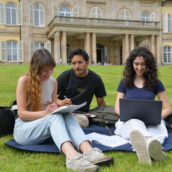 Foto: Drei Personen sitzen auf einer Wiese vor Schloss Hohenheim. Eine Person schreibt in ein Notizbuch, eine andere arbeitet an einem Laptop. Im Hintergrund ist das Schloss mit seinen Säulen und Fenstern sichtbar.