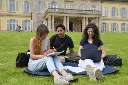 Foto: Drei Personen sitzen auf einer Wiese vor Schloss Hohenheim. Eine Person schreibt in ein Notizbuch, eine andere arbeitet an einem Laptop. Im Hintergrund ist das Schloss mit seinen Säulen und Fenstern sichtbar.