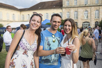 Foto: Drei Personen stehen vor Schloss Hohenheim. Sie halten Getränke in der Hand. Im Hintergrund sind weitere Personen und das Schloss sichtbar.