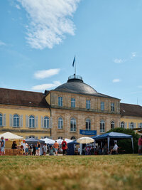 Foto: Schloss Hohenheim mit mehreren Marktständen und Personen im Vordergrund. Ein blauer Himmel mit einigen Wolken ist sichtbar. Ein Banner mit der Aufschrift 'Universität Hohenheim' hängt an einem Stand.