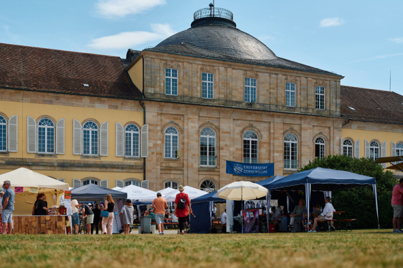 Foto: Schloss Hohenheim mit mehreren Marktständen und Personen im Vordergrund. Ein blauer Himmel mit einigen Wolken ist sichtbar. Ein Banner mit der Aufschrift 'Universität Hohenheim' hängt an einem Stand.